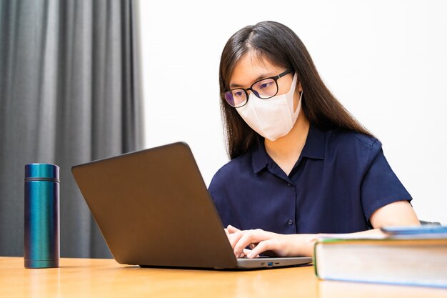 Joven asiática con máscara N95, trabajando en su computadora desde casa durante la propagación del virus COVID-19