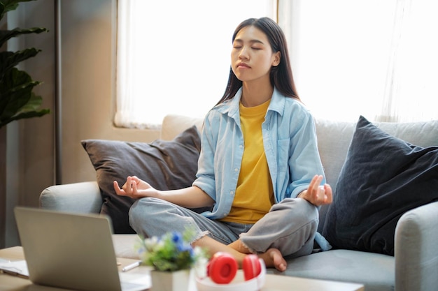 Joven asiática meditando mientras se sienta en el sofá en casa