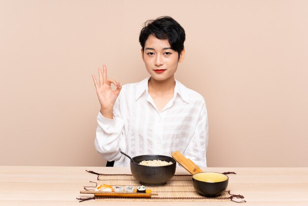 Joven asiática en una mesa con tazón de fideos y sushi mostrando un signo bien con los dedos