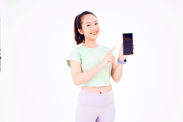 La joven asiática muestra una pantalla de teléfono inteligente vacía con expresión positiva sonríe ampliamente vestida con ropa informal sintiendo felicidad en el fondo azul Teléfono móvil con pantalla blanca en la mano femenina