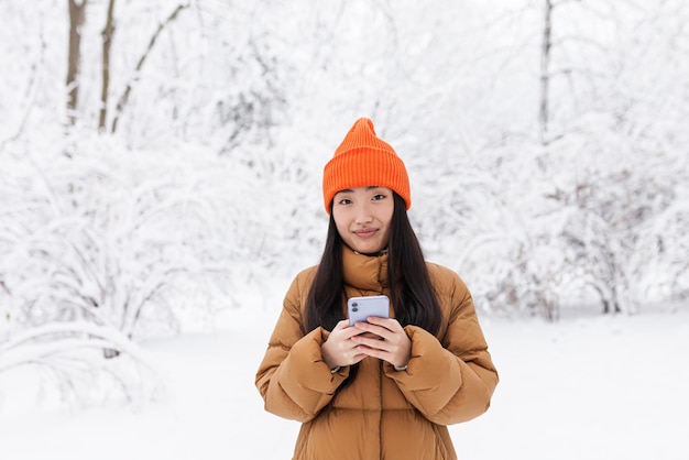 Joven asiática, en el parque en una cita, esperando, en un día nevado de invierno, en línea usa el teléfono