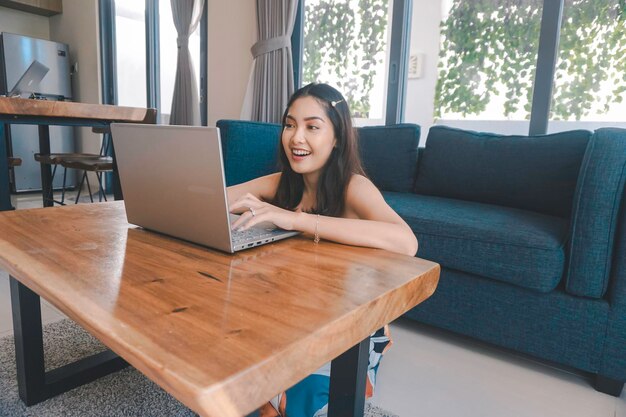 La joven asiática pasa su tiempo en casa sentada en la sala de estar sonriendo y trabajando en su computadora portátil