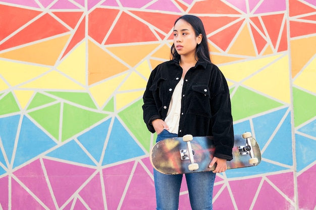 Joven asiática con una patineta en la mano se encuentra frente a una pared colorida