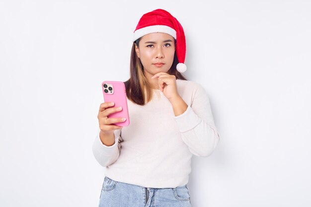Una joven asiática pensativa con un sombrero de Navidad sosteniendo un teléfono móvil y tocándose la barbilla aislada sobre el fondo blanco