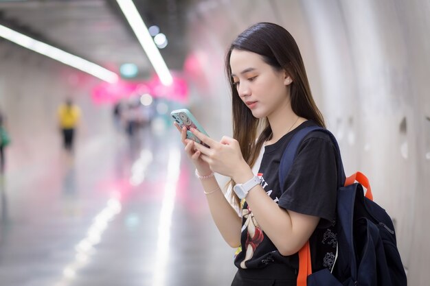 Joven asiática que tiene el pelo largo tiene un teléfono inteligente en la mano y se encuentra en el túnel del metro