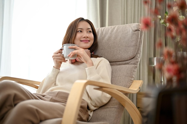 Una joven asiática relajada y relajada está recostada en su sillón tomando un café por la mañana