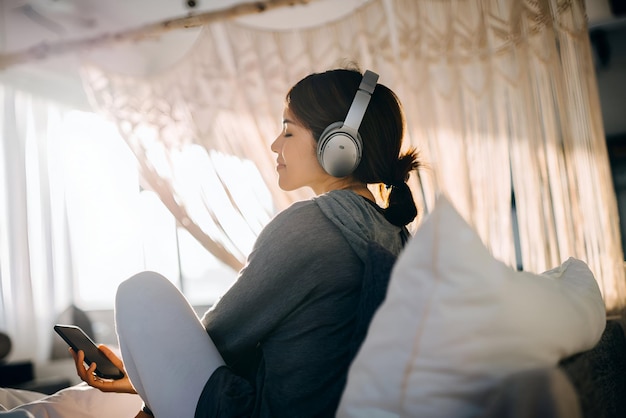 Una joven asiática relajada sentada en su cama disfrutando de música en los auriculares de su teléfono inteligente en casa