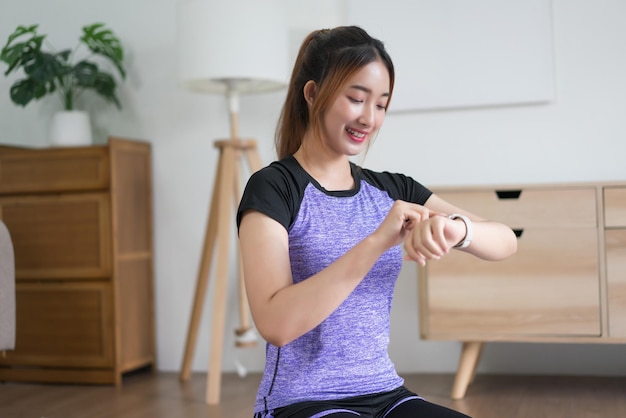 Joven asiática revisando el tiempo de entrenamiento y la frecuencia cardíaca en el reloj de pulsera después de hacer ejercicio en casa