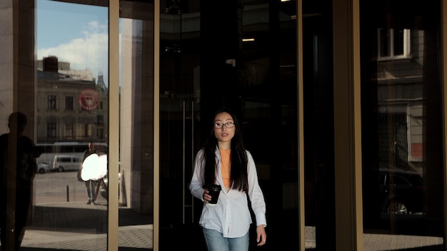 Joven asiática saliendo del edificio moderno y tomando café al aire libre retrato