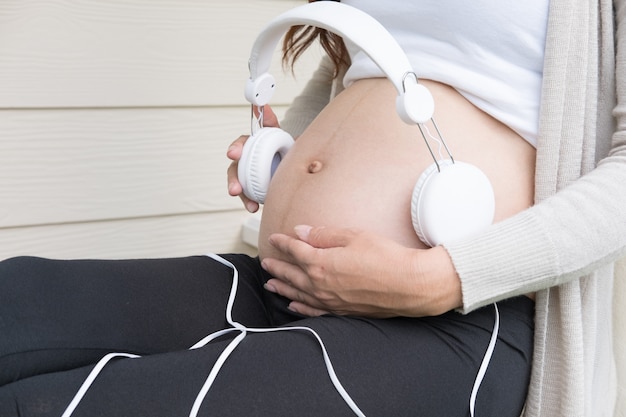 Joven asiática sentada en el balcón con auriculares en su vientre tocando música a un bebé en el útero.