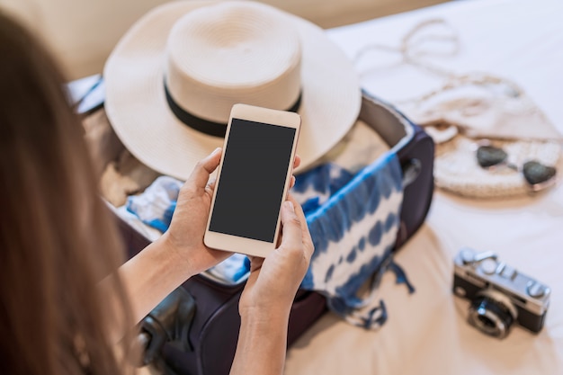 Foto joven asiática sentada en la cama con smartphone y empacando su maleta preparándose para viajar en vacaciones de verano