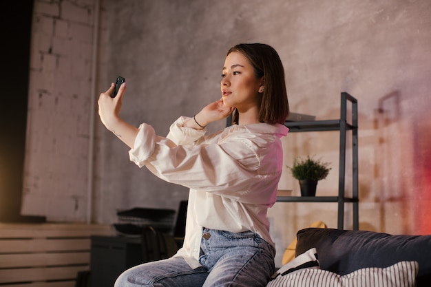 Foto joven asiática sentada en un sofá en una acogedora habitación toma un selfie y sonríe