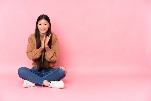 Foto joven asiática sentada en el suelo en la pared rosa aplaudiendo después de la presentación en una conferencia