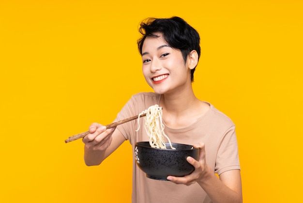 Joven asiática sobre pared amarilla aislada sosteniendo un tazón de fideos con palillos y ofreciéndolo