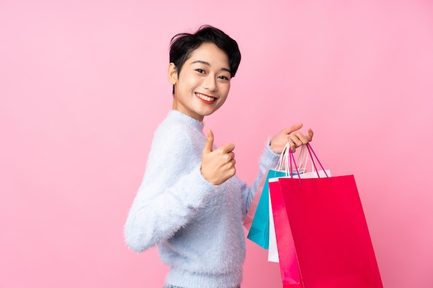 Joven asiática sobre pared rosa aislada con bolsas de compras y con el pulgar hacia arriba