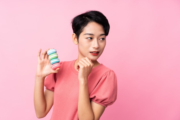 Joven asiática sobre pared rosa aislado con coloridos macarons franceses y pensando en una idea