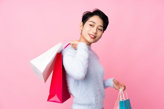 Joven asiática sobre pared rosa aislado sosteniendo bolsas de compras y sonriendo