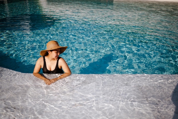 Joven asiática con sombrero relajándose junto a la piscina en un hotel de lujo y concepto de verano