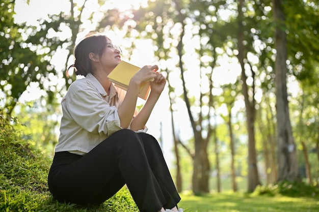 Una joven asiática soñadora e inspirada se sienta en el césped en el parque verde con un libro