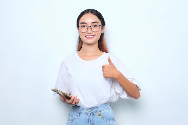 Una joven asiática sonriente con camiseta blanca usando un smartphone y mostrando el pulgar hacia arriba aislada de fondo blanco