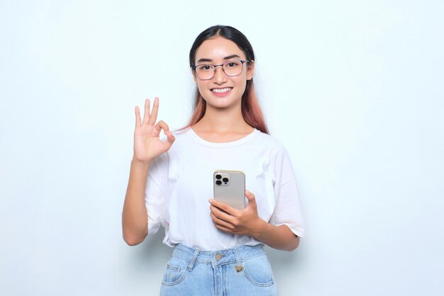 Una joven asiática sonriente con camiseta blanca usando un teléfono inteligente y mostrando un gesto correcto aislado de fondo blanco