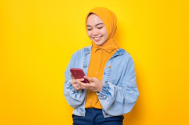 Una joven asiática sonriente con chaqueta de jeans usando un teléfono móvil escribiendo mensajes SMS aislados en un fondo amarillo