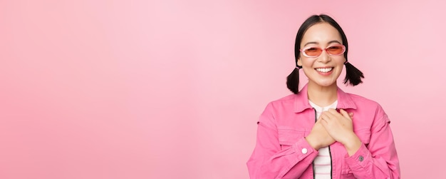 Una joven asiática sonriente con gafas de sol que parece agradecida cogida de la mano en el corazón halagada aprecia algo de pie sobre un fondo rosa