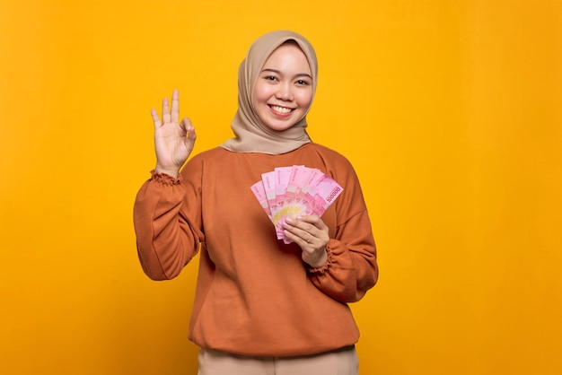 Una joven asiática sonriente con pantalones naranja sosteniendo billetes de dinero y haciendo un gesto de aprobación aislado sobre el fondo amarillo