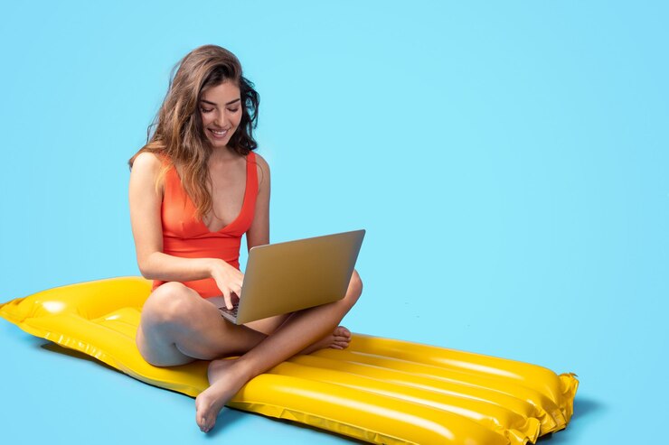 Una joven asiática sonriente en traje de baño se sienta en un colchón inflable con una computadora portátil y disfruta del trabajo y la natación