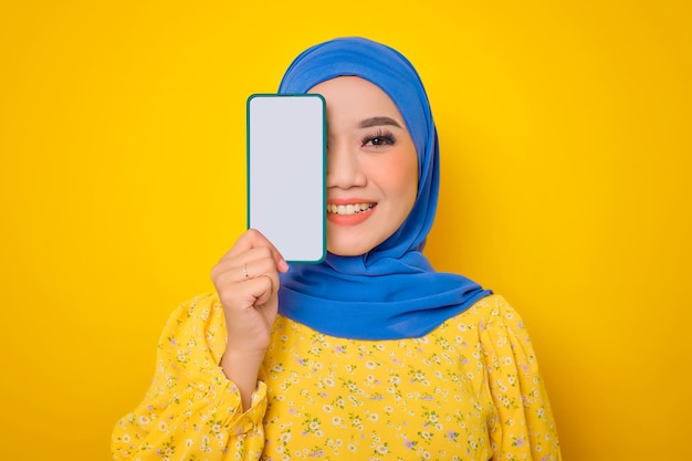 Una joven asiática sonriente vestida de forma informal que cubre la mitad de la cara con un teléfono móvil con pantalla en blanco recomendando una aplicación móvil aislada de fondo amarillo