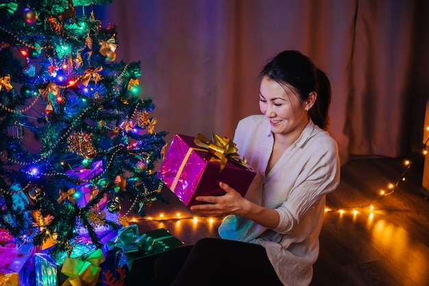 Una joven asiática con una sonrisa en su rostro tiene en sus manos una caja de regalo de Navidad de color morado con un lazo amarillo. Fondo de Navidad con árbol de Navidad, guirnaldas y regalos.