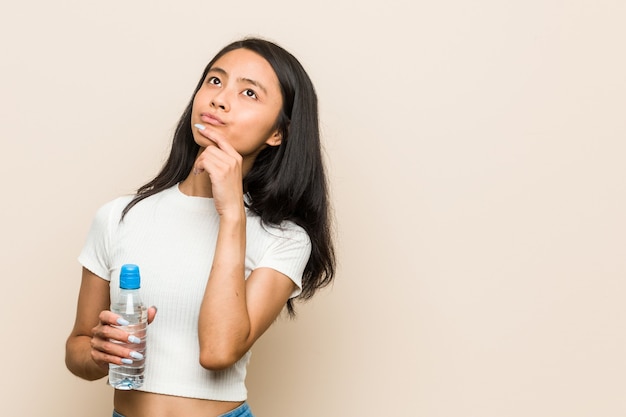 Joven asiática sosteniendo una botella de agua mirando hacia los lados con expresión dudosa y escéptica.