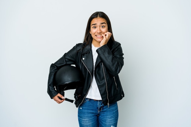 Joven asiática sosteniendo un casco de moto sobre pared aislada mordiéndose las uñas, nerviosa y muy ansiosa