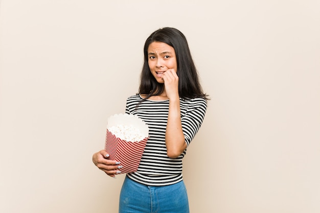 Joven asiática sosteniendo un cubo de palomitas de maíz mordiendo las uñas, nervioso y muy ansioso.