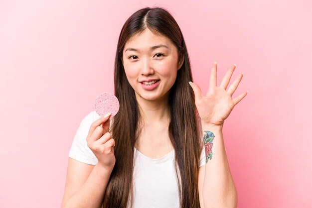 Joven asiática sosteniendo una esponja facial aislada en un fondo rosa sonriendo alegre mostrando el número cinco con los dedos