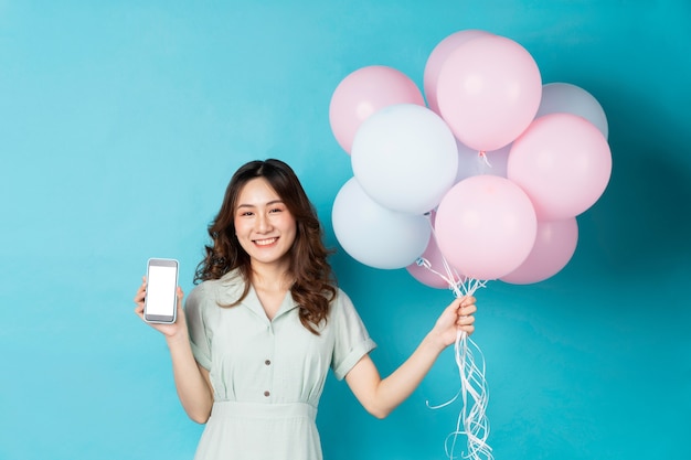 Joven asiática sosteniendo globos y usando el teléfono con expresión feliz en turquesa