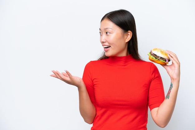 Joven asiática sosteniendo una hamburguesa aislada de fondo blanco con expresión facial sorpresa