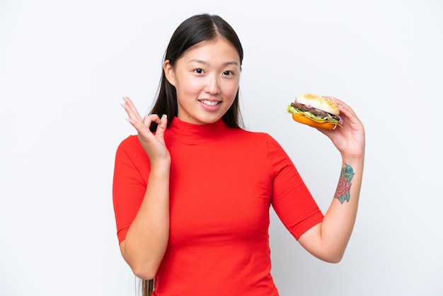 Joven asiática sosteniendo una hamburguesa aislada de fondo blanco mostrando el signo de ok con los dedos