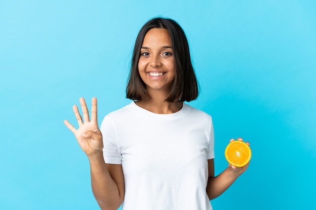 Joven asiática sosteniendo una naranja aislada en la pared azul feliz y contando cuatro con los dedos