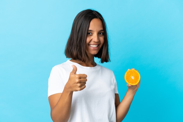 Joven asiática sosteniendo una naranja aislada en la pared azul con los pulgares hacia arriba porque ha sucedido algo bueno