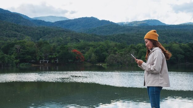 Una joven asiática sosteniendo y usando un teléfono móvil mientras viaja por las montañas y el lago