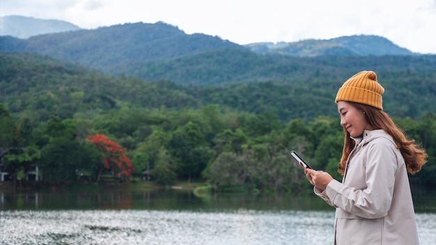 Una joven asiática sosteniendo y usando un teléfono móvil mientras viaja por las montañas y el lago