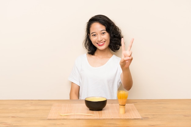 Joven asiática con té matcha sonriendo y mostrando el signo de la victoria