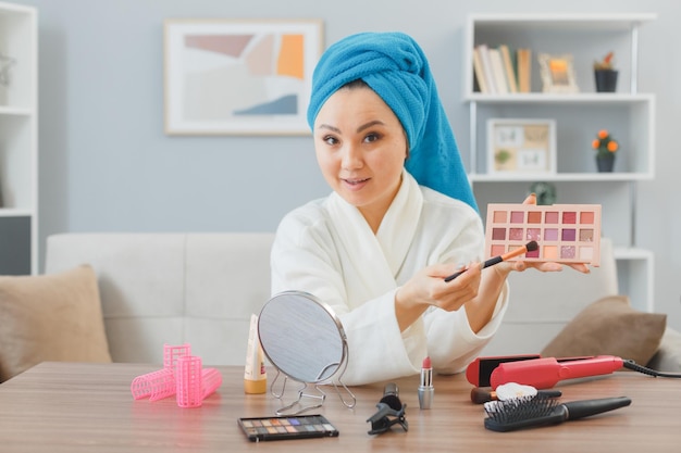 Joven asiática con una toalla en la cabeza sentada en el tocador en el interior de su casa aplicando sombras de ojos sosteniendo una paleta feliz y positiva haciendo belleza de rutina de maquillaje matutino