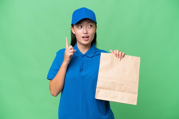 Joven asiática tomando una bolsa de comida para llevar sobre un fondo aislado pensando en una idea apuntando con el dedo hacia arriba