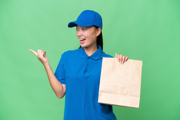 Joven asiática tomando una bolsa de comida para llevar sobre un fondo aislado señalando con el dedo al costado y presentando un producto