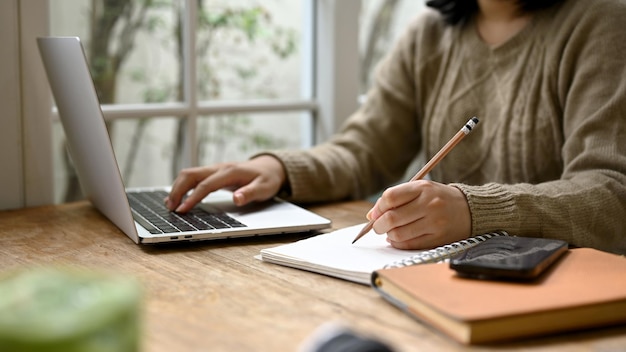 Una joven asiática tomando notas en su cuaderno y usando su laptop en una cafetería