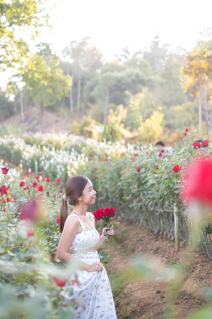 Una joven asiática con un vestido blanco posa con una rosa en un jardín de rosas