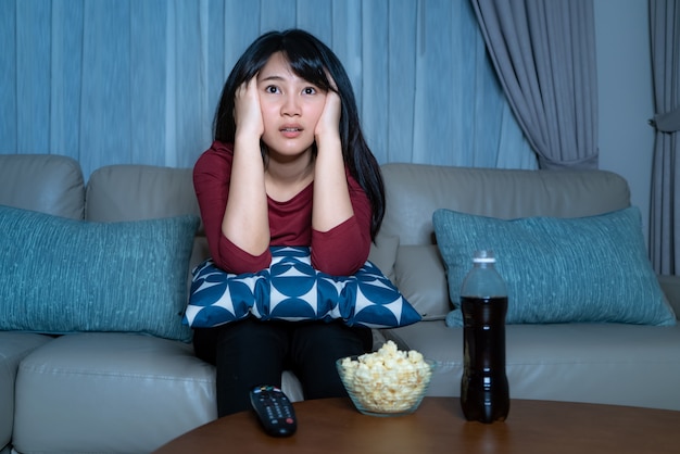 Joven asiática viendo televisión suspenso película o noticias mirando sorprendido y emocionado comiendo palomitas de maíz tarde en la noche en casa sofá de la sala durante el tiempo de aislamiento en el hogar