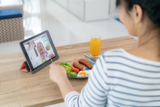 Foto joven asiática virtual happy hour reuniéndose y comiendo comida en línea junto con su madre en video conferencia con tableta digital para una reunión en línea en video llamada